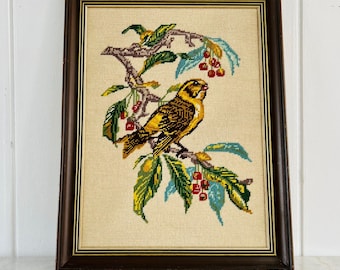 May include: A framed needlepoint embroidery featuring a yellow and brown bird perched on a branch with green leaves and small red berries. The artwork is set within a dark brown wooden frame with gold inner trim, displayed on a light background.