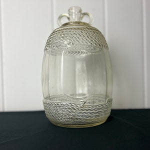 May include: A clear glass bottle shaped like a barrel with a rope design. The bottle has a cork stopper and two handles.
