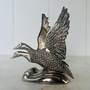 May include: A silver-colored sculpture of two ducks in flight. The sculpture has detailed feather and wing textures, with one duck's wings fully extended. The ducks are mounted on a stylized wave base, creating a dynamic and elegant decorative piece.