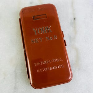 Puede incluir: Un estuche metálico rectangular de color óxido vintage con el texto en relieve "YORK SET No. 9 MATHEMATICAL INSTRUMENTS". El estuche tiene una tapa con bisagras y una pequeña abertura rectangular en la parte superior.