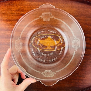 Peut inclure: Un bol en verre transparent avec un motif doré représentant une lampe surmontée d'une couronne. Le bol a un bord surélevé et une petite poignée de chaque côté. Le bol présente un motif de petits trous autour du fond.