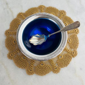 Puede incluir: Un cuenco plateado lleno de líquido azul oscuro, con una cuchara plateada decorativa en su interior. El cuenco descansa sobre un tapete de ganchillo dorado. La cuchara tiene un diseño en forma de concha.