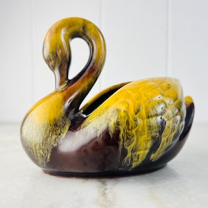 May include: A ceramic swan-shaped planter with a glossy finish. The planter has a curved neck and a wide opening for plants. The colour scheme includes shades of brown, yellow, and black, creating a striking visual contrast. The swan is a decorative item.