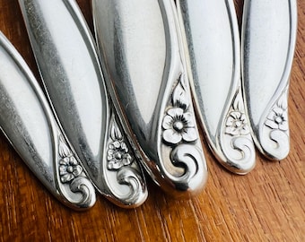 May include: Close-up of several silver-colored utensils, possibly silverware, with a floral design at the handle's base. The utensils are arranged on a wooden surface, showcasing their reflective quality and intricate detailing.