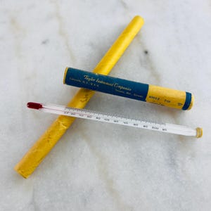 May include: A vintage Taylor Instrument Companies thermometer with a red bulb, marked with degree increments. The thermometer is displayed with its original yellow and blue paper packaging. The packaging includes text from Asheville, N.C., U.S.A. and Toronto, Ont., Canada.