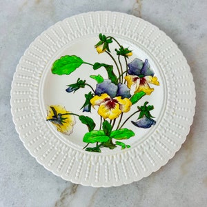 Puede incluir: Plato de cerámica blanco con un borde de cuentas en relieve. El centro del plato presenta un diseño pintado a mano de pensamientos amarillos y morados con hojas verdes.