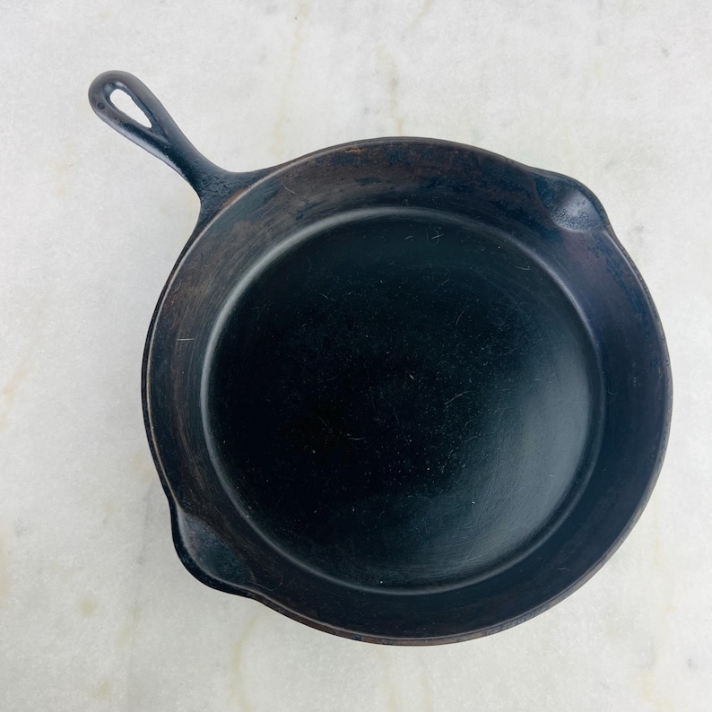 May include: A well-seasoned, black cast iron skillet with a handle and a small hole for hanging. The skillet has a round cooking surface and a slightly raised edge. The pan is designed for stovetop or oven use.