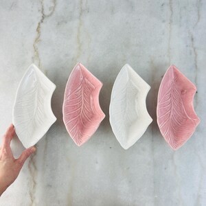 May include: Four leaf-shaped ceramic dishes, two white and two pink, arranged on a marble surface. Each dish features a detailed leaf vein pattern. A hand is holding one of the white dishes.