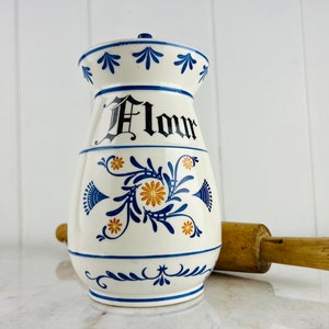 May include: White ceramic flour canister with blue and yellow floral designs and the word "Flour" in black lettering. The canister is sitting on a white surface next to a wooden rolling pin.