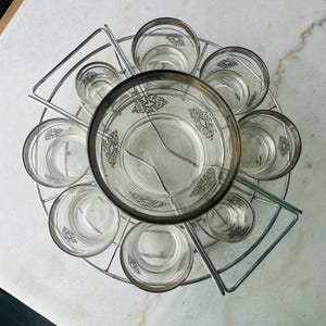 May include: A vintage glassware set with a central bowl and eight glasses, each with silver rims and etched patterns. The glasses are held in a circular metal rack. Ideal for serving drinks at a gathering.