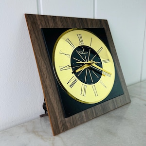 May include: A vintage square clock with a gold-coloured face and black Roman numerals. The clock face is set against a black background, framed by a wood-grain border. The clock is angled, showing its side profile.