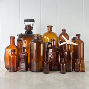 May include: An assortment of amber glass bottles in different shapes and sizes. Some bottles have labels, others are plain. A few have stoppers or lids. A white starfish sits atop one bottle. The bottles are arranged on a light-coloured surface.
