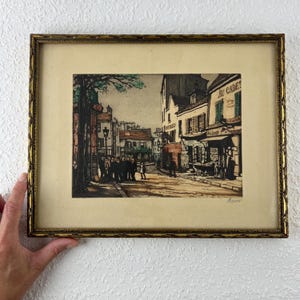 May include: Framed vintage print depicting a street scene with buildings, people, and the text "Au Cadet". The print is in a gold ornate frame. The artwork features a muted color palette with shades of brown, beige, and green.