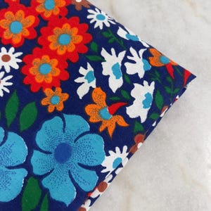 May include: Folded fabric with a vibrant floral pattern. The design features large blue, orange, and white flowers on a dark blue background. Green leaves and brown accents complete the colorful print, creating a retro aesthetic.
