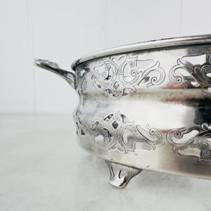 Puede incluir: Plato de servir de plata con un diseño decorativo de corte y tres patas. El plato tiene un asa en un lado.