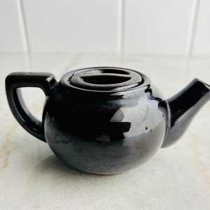 May include: A small black ceramic teapot with a lid. The teapot has a rounded body and a spout on the side.
