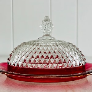 Può includere: Un porta burro vintage in vetro con un coperchio trasparente a forma di diamante e una base rosso rubino.