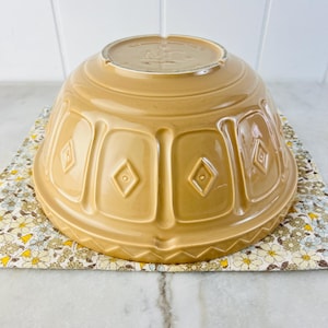 May include: A tan ceramic mixing bowl with a decorative diamond pattern. The bowl is upside down, revealing its base. It rests on a floral patterned cloth. The bowl is a warm, neutral color, suitable for kitchen use.