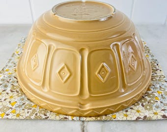 Puede incluir: Un cuenco de cerámica beige con un diseño de diamantes. El cuenco está boca abajo, revelando su base. Descansa sobre una tela con estampado floral. El cuenco es de un color cálido y neutro, adecuado para la cocina.