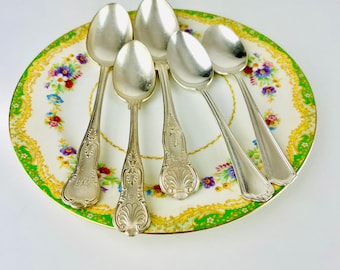 May include: Five silver spoons with ornate handles are arranged on a green and white floral patterned plate.