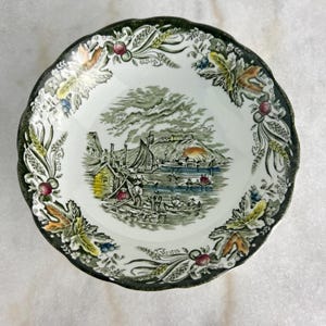 May include: A vintage green and white ceramic bowl with a detailed scene of a harbor town with boats and people. The bowl has a scalloped edge and a green and gold floral design.
