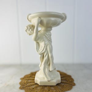 Op de afbeelding: Crèmekleurig keramieken beeld van een figuur die een grote kom vasthoudt. De figuur is gehuld in gewaden en staat op een vierkante basis. Het beeld staat op een bruin, gehaakt kleedje.