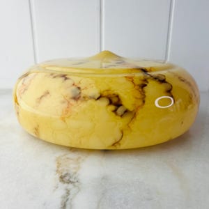 May include: A yellow and brown marbled glass bowl with a lid. The bowl has a rounded shape with a slightly pointed top on the lid. The colors are a mix of yellow, brown, and red, creating a unique pattern. The bowl is sitting on a white surface.