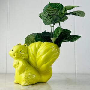 May include: A bright yellow ceramic squirrel planter holding green artificial leaves. The squirrel is in a sitting position with its tail curled up. The planter is a decorative item for home decor.