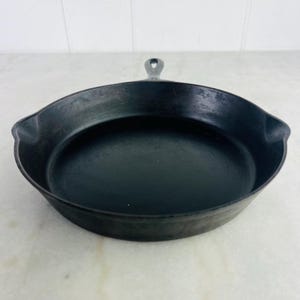 May include: A black cast iron skillet with a single handle. The skillet is round with a slightly raised edge and a dark, non-reflective surface. The handle has a hole for hanging.