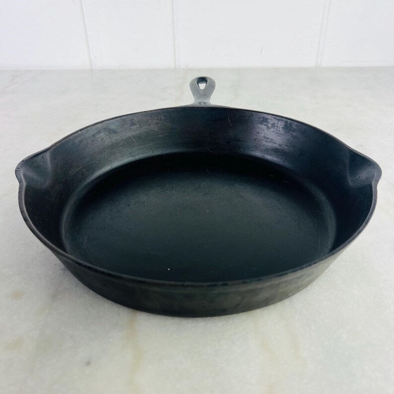 May include: A black cast iron skillet with a single handle. The skillet is round with a slightly raised edge and a dark, non-reflective surface. The handle has a hole for hanging.