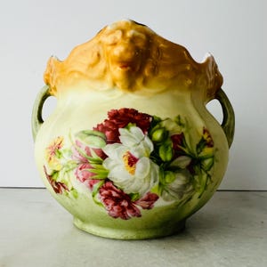 May include: A ceramic vase featuring a pale yellow and green body with a hand-painted floral arrangement of white, red, and pink blossoms. The rim is sculpted with a golden-brown lion head motif, and the vessel includes two green side handles.