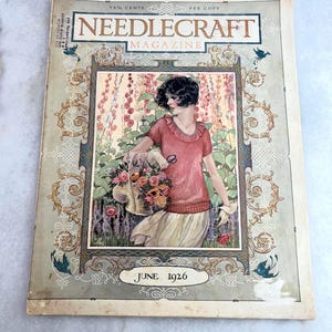 Może przedstawiać: Okładka magazynu "Needlecraft" z czerwca 1926 roku. Na okładce znajduje się ilustracja kobiety w różowym topie i spódnicy trzymającej kosz kwiatów. Magazyn ma dekoracyjne obramowania i tekst "Needlecraft Magazine" u góry.