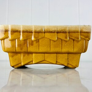 May include: A yellow ceramic planter with a basket weave design. The planter has a rectangular shape with a slightly rounded bottom and a raised lip around the top.