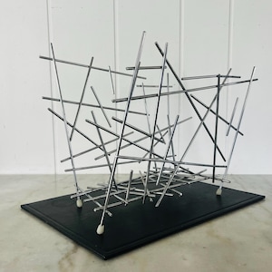May include: A chrome metal magazine rack featuring a series of intersecting thin rods arranged in a chaotic, linear pattern. The structure is supported by a rectangular black base with small white protective feet at each corner of the metal frame.