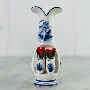 Puede incluir: Un jarrón de cerámica blanco con un diseño de molino de viento azul y blanco y la palabra "Holland". El jarrón presenta dos zuecos en miniatura con flores rojas. El jarrón mide aproximadamente 18 cm de alto.
