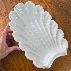 May include: White ceramic seashell dish with a textured surface. The dish is shaped like a large seashell with a rounded edge.