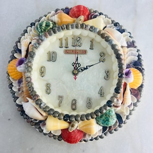 Peut inclure: Une horloge murale ronde ornée de divers coquillages blancs, beiges, rouges, jaunes et verts. Le cadran est blanc avec des chiffres dorés et des aiguilles noires. Le mot "PHILIPPINES" est imprimé sur le cadran.