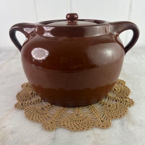 Può includere: Una pentola in ceramica marrone con coperchio e due manici. La pentola è appoggiata su un sottobicchiere all'uncinetto color oro. La pentola ha una finitura lucida e un piccolo pomello sul coperchio.