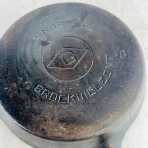 May include: Close-up of a well-worn, cast iron skillet. The skillet's base features the embossed text "SMART" above a diamond-shaped logo with a "G" inside, and "BROCKVILLE, PA" below. The pan has a dark, aged appearance.