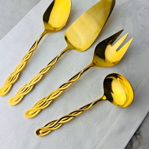 May include: A set of four gold-colored serving utensils. The set includes a spoon, cake server, fork, and ladle. Each utensil has a twisted handle design. The items are arranged on a white, patterned cloth.