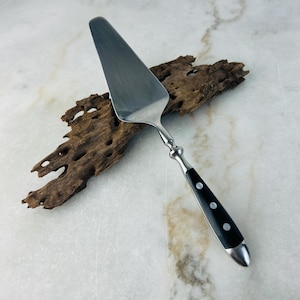 May include: A stainless steel cake server with a black handle featuring three white dots. The server has a silver-coloured handle and a wide, triangular blade. The server is displayed on a piece of driftwood.