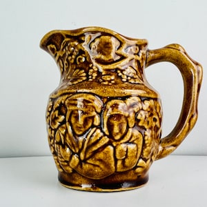 May include: A brown ceramic pitcher with a detailed embossed design of people and grapes. The pitcher has a handle and a rounded body.