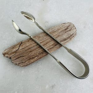 May include: Silver-toned sugar tongs with spoon-shaped ends, resting on a piece of driftwood. The tongs have a curved handle for easy gripping. The background is a light-colored, textured surface.