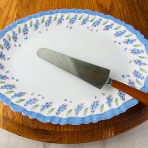 Könnte beinhalten: Ovale Servierplatte mit einem gewellten blauen Rand und floralem Design. Ein Kuchenheber aus Edelstahl mit Holzgriff liegt auf der Platte. Die Platte ist weiß mit blauen und grünen floralen Akzenten.