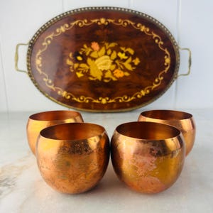 May include: An oval wooden serving tray with a floral inlay design and brass handles, alongside four copper-coloured, round, bowl-shaped cups. The tray has a dark brown background with a decorative border.