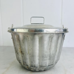 Op de afbeelding: Een vintage, gegolfde metalen tulbandvorm met een deksel. De vorm is zilverkleurig met een handvat op het deksel en kleine handvatten aan de zijkanten. De vorm heeft een decoratief, geribbeld ontwerp en vertoont tekenen van ouderdom.