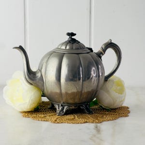 Puede incluir: Una tetera de plata con un cuerpo redondeado en forma de calabaza y un diseño floral en la tapa. La tetera tiene un asa decorativa y se apoya en cuatro patas.