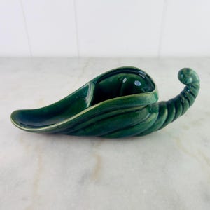 May include: A dark green ceramic cornucopia-shaped dish. The dish has a glossy finish and a curved, horn-like shape with a textured, ribbed design on the curled end. The interior has a smooth, open space for display.