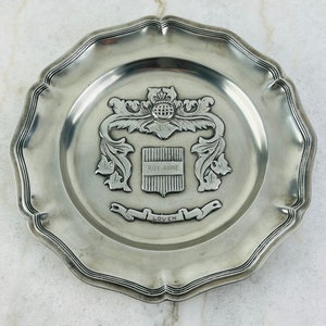 May include: A silver-toned metal plate with a decorative crest featuring a coat of arms with the text "ROY-AINE" and "LOVEN".