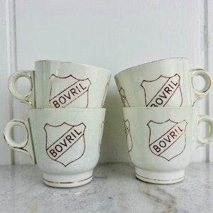 May include: Four off-white ceramic cups with handles, stacked in two pairs. Each cup features a shield design with the word "BOVRIL" in a dark red colour. The rims and handles are trimmed with gold.